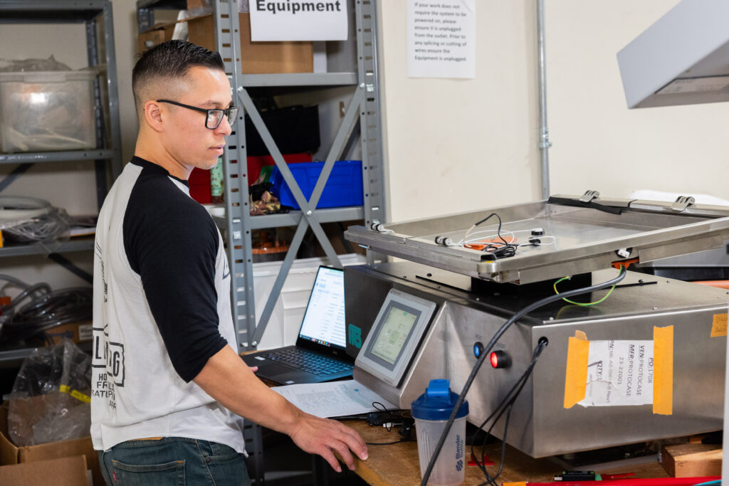 A focused technician in a workshop operates a high-tech industrial machine, monitoring data on a touchscreen interface and a laptop. The workspace is equipped with shelves holding various tools and equipment, reflecting a setting of precision engineering and testing.