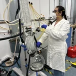 Scientist wearing protective gear operates a rotary evaporator in a laboratory setting. The setup includes glass condensers, tubing, and a round-bottom flask, highlighting a professional lab environment focused on chemical or pharmaceutical processes.