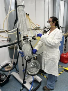 Scientist wearing protective gear operates a rotary evaporator in a laboratory setting. The setup includes glass condensers, tubing, and a round-bottom flask, highlighting a professional lab environment focused on chemical or pharmaceutical processes.