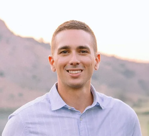 Headshot of Christopher VanBuskirk, smiling outdoors in a light blue collared shirt with a blurred mountain background.